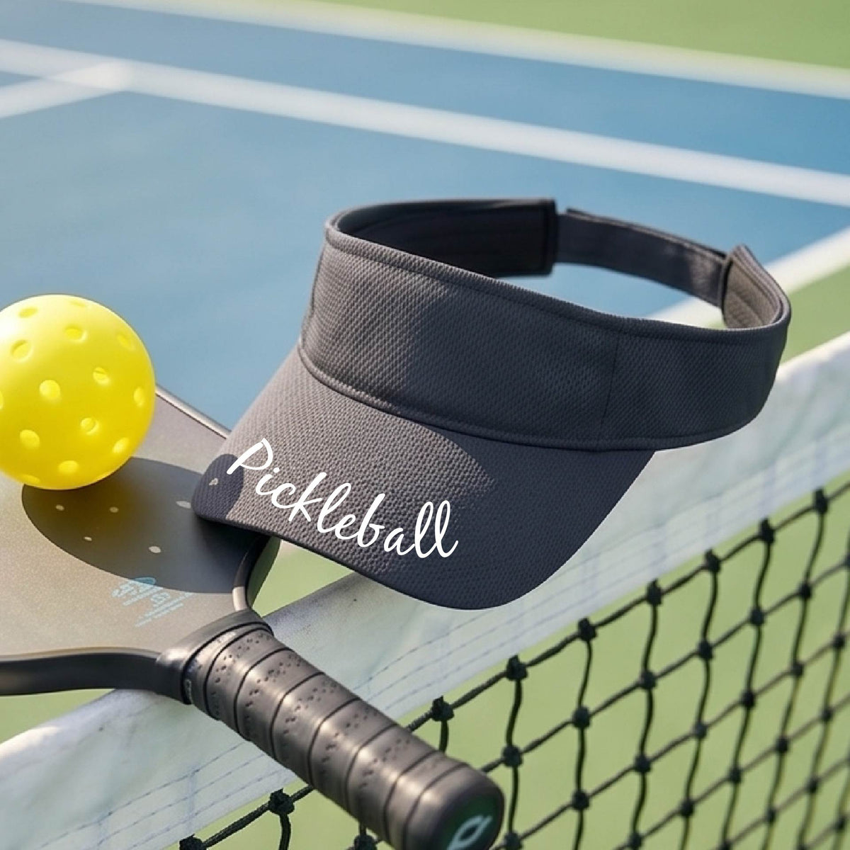A close-up of a graphite gray visor with a graphic of the word “Pickleball” in a white script across the brim of the visor, shown worn in front of a pickleball court in the background. 