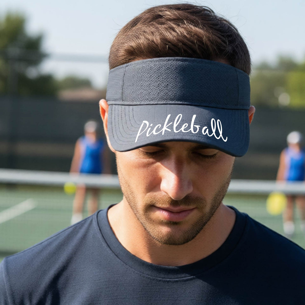 A close-up of a graphite gray visor with a graphic of the word “Pickleball” in a white script across the brim of the visor, shown worn in front of a pickleball court in the background. 