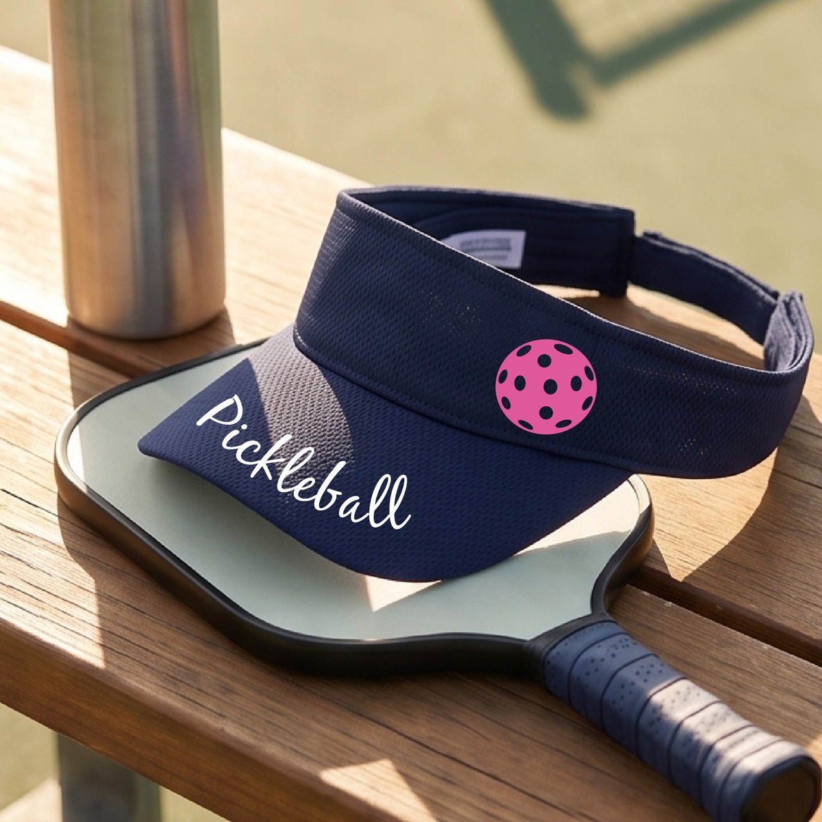 A close-up of a navy blue visor with a graphic of the word “Pickleball” in a white script across the brim of the visor, including a colorful pickleball shown worn in front of a pickleball court in the background. 
