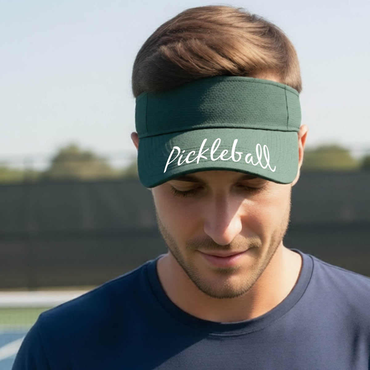 A close-up of a green visor with a graphic of the word “Pickleball” in a white script across the brim of the visor, shown worn in front of a pickleball court in the background. 