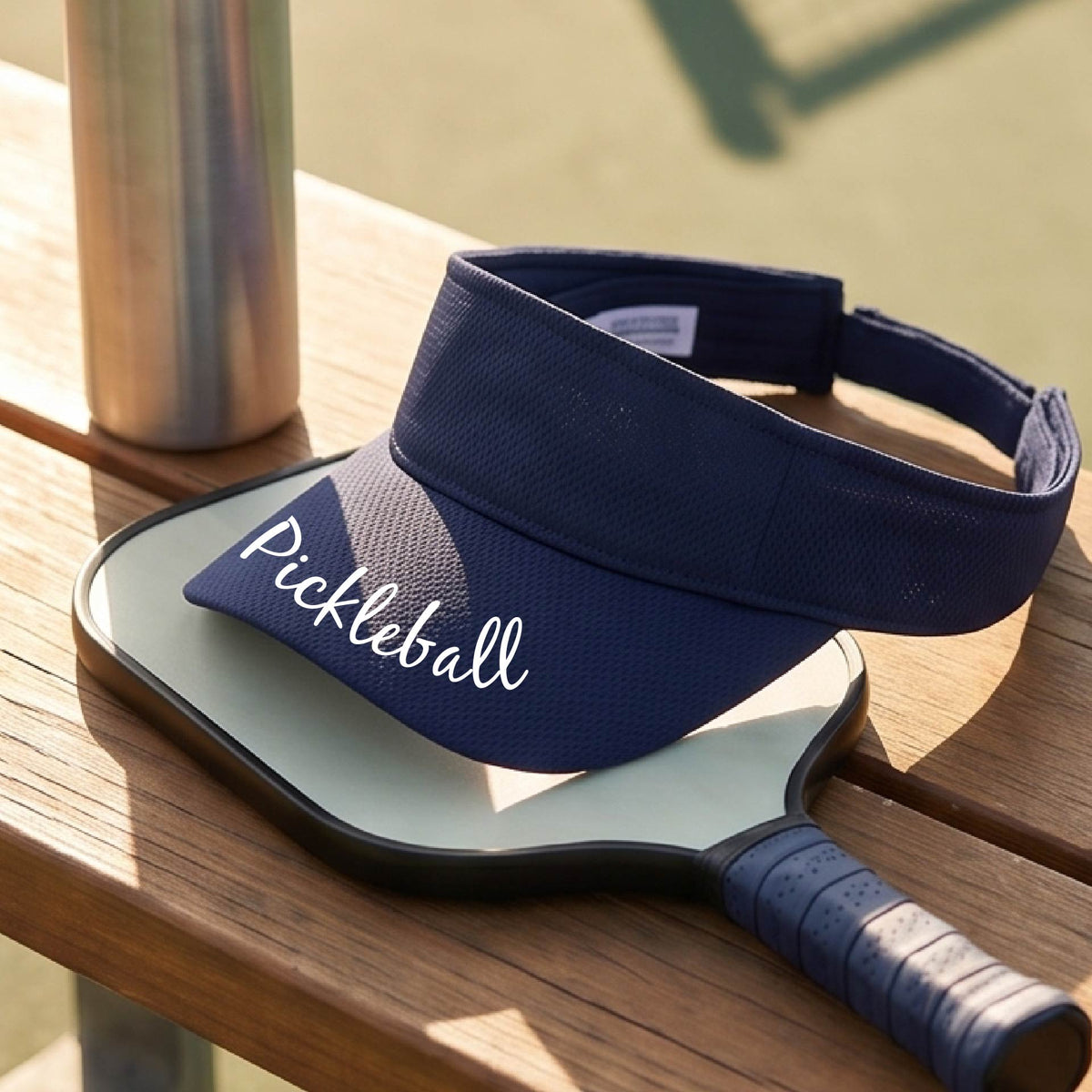 A close-up of a navy blue visor with a graphic of the word “Pickleball” in a white script across the brim of the visor, shown worn in front of a pickleball court in the background. 