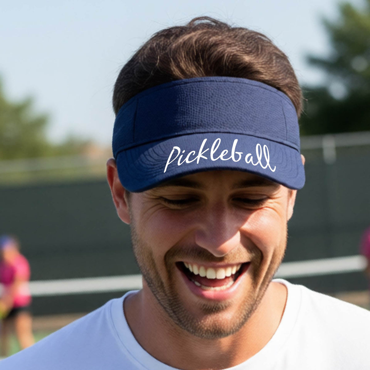 A close-up of a navy blue visor with a graphic of the word “Pickleball” in a white script across the brim of the visor, shown worn in front of a pickleball court in the background. 