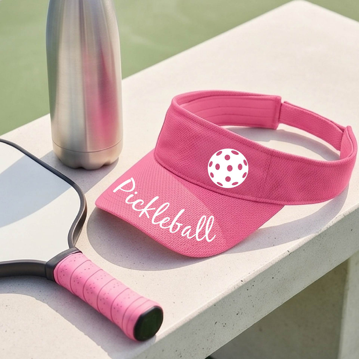 A close-up of a pink visor with a graphic of the word “Pickleball” in a white script across the brim of the visor, including a colorful pickleball shown worn in front of a pickleball court in the background. 