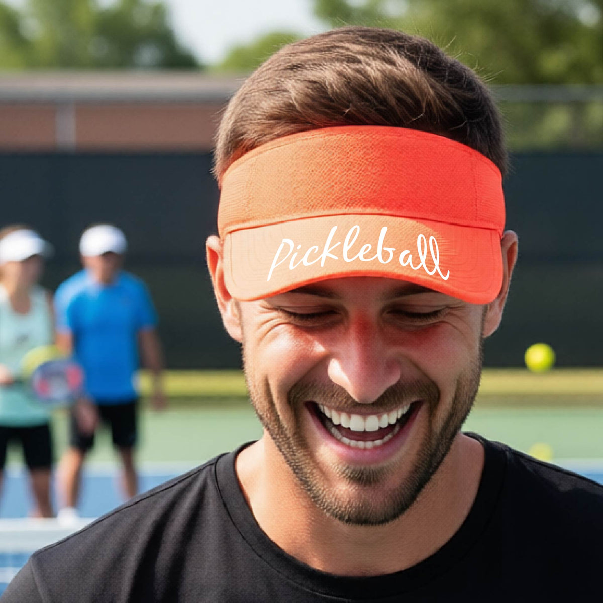 A close-up of an orange visor with a graphic of the word “Pickleball” in a white script across the brim of the visor, shown worn in front of a pickleball court in the background. 