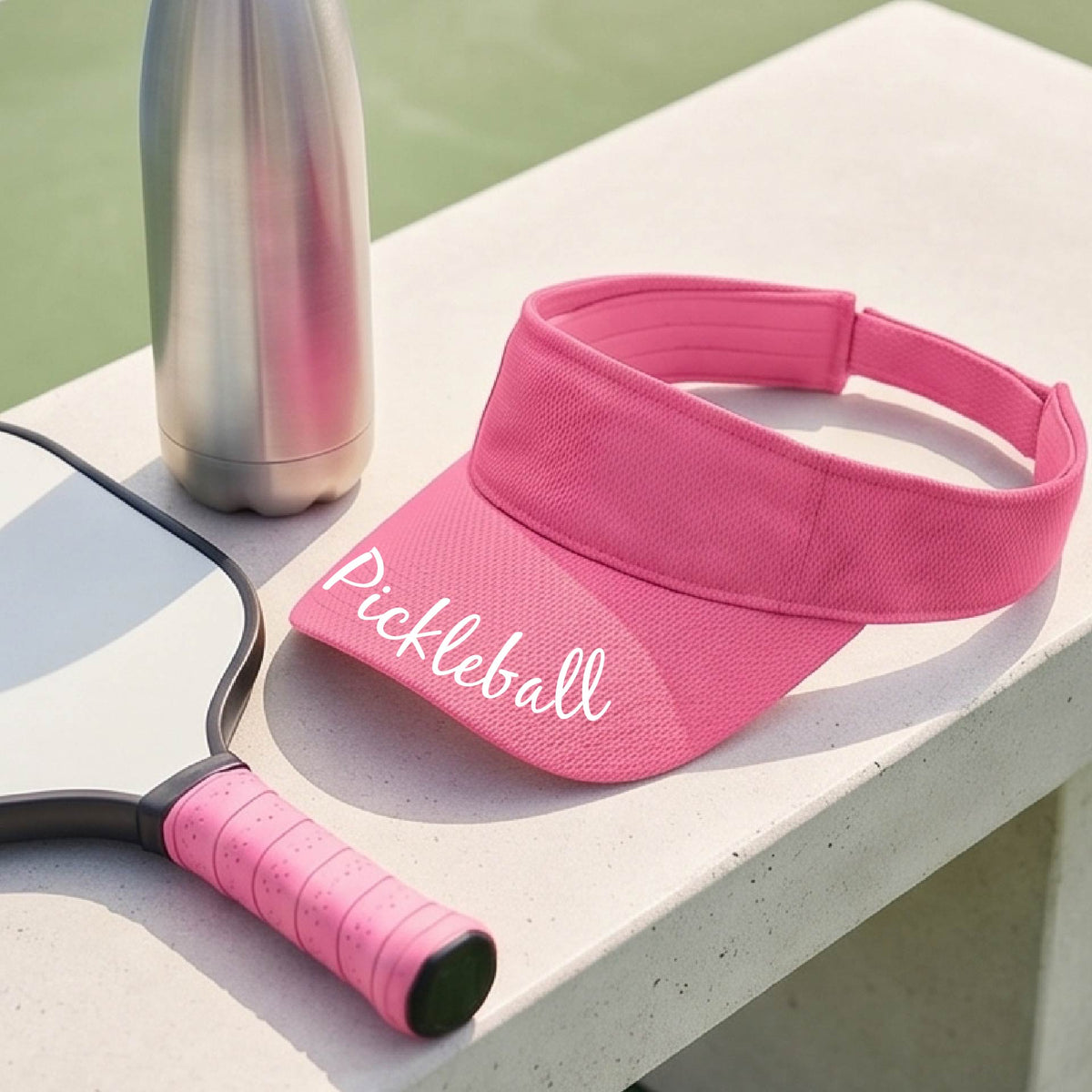 A close-up of a pink visor with a graphic of the word “Pickleball” in a white script across the brim of the visor, shown worn in front of a pickleball court in the background. 