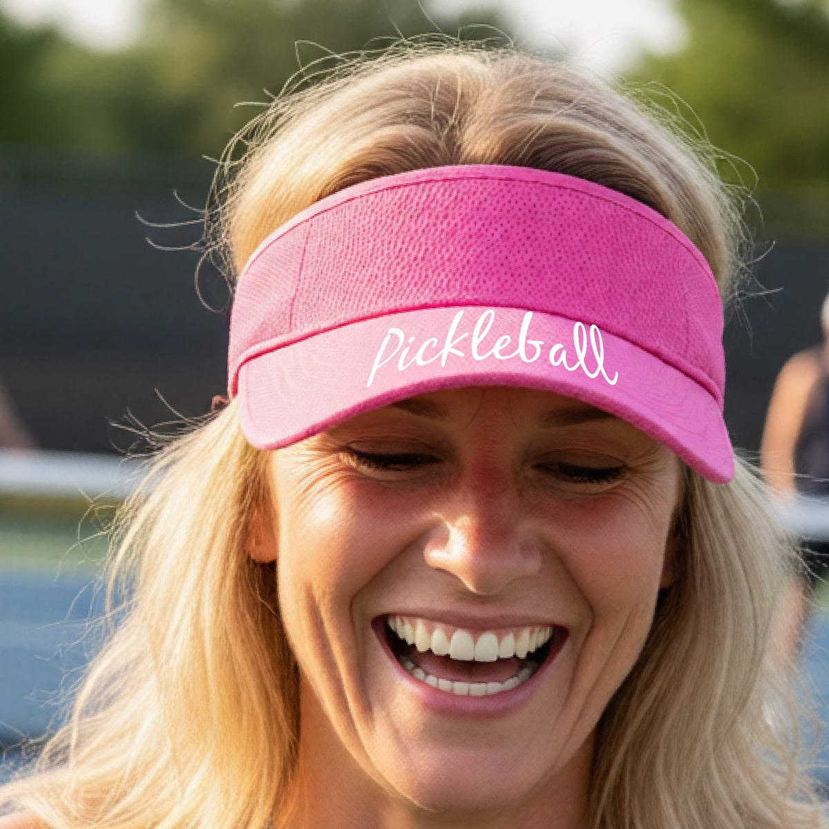 A close-up of a pink visor with a graphic of the word “Pickleball” in a white script across the brim of the visor, shown worn in front of a pickleball court in the background. 