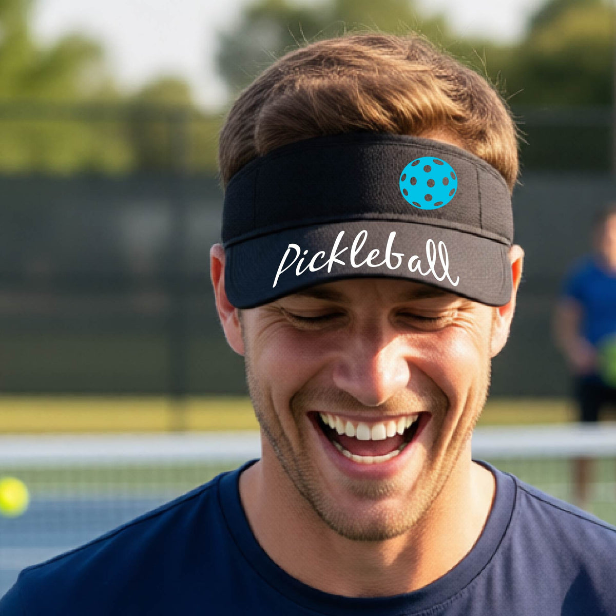 A close-up of a black visor with a graphic of the word “Pickleball” in a white script across the brim of the visor, including a colorful pickleball shown worn in front of a pickleball court in the background. 