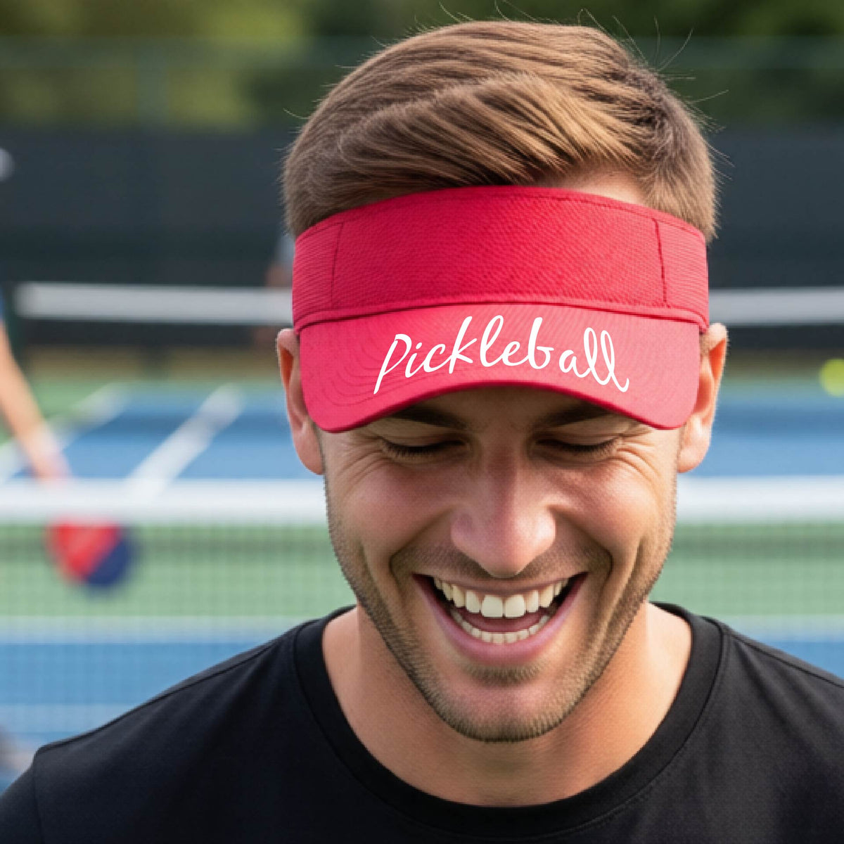 A close-up of a red visor with a graphic of the word “Pickleball” in a white script across the brim of the visor, shown worn in front of a pickleball court in the background. 