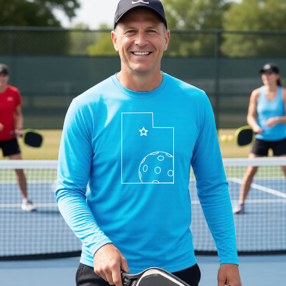 A close-up of a man wearing a bright blue short-sleeve t-shirt, with a white graphic outline of the State of Utah , including an outline of a white pickleball in the lower right corner and a star outline in the center of the graphic, on the back of the shirt.   