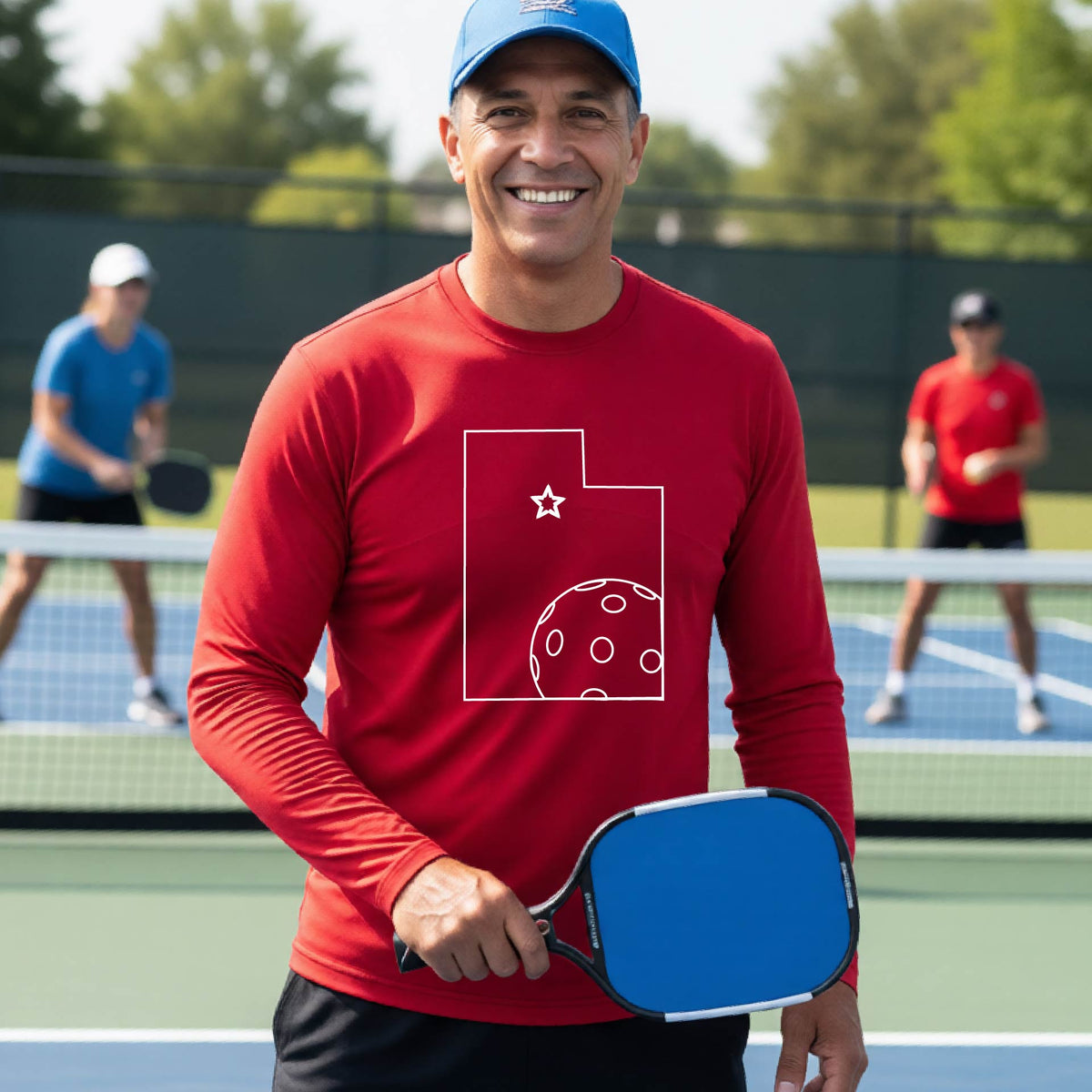 A close-up of a man wearing a red short-sleeve t-shirt, with a white graphic outline of the State of Utah , including an outline of a white pickleball in the lower right corner and a star outline in the center of the graphic, on the back of the shirt.   
