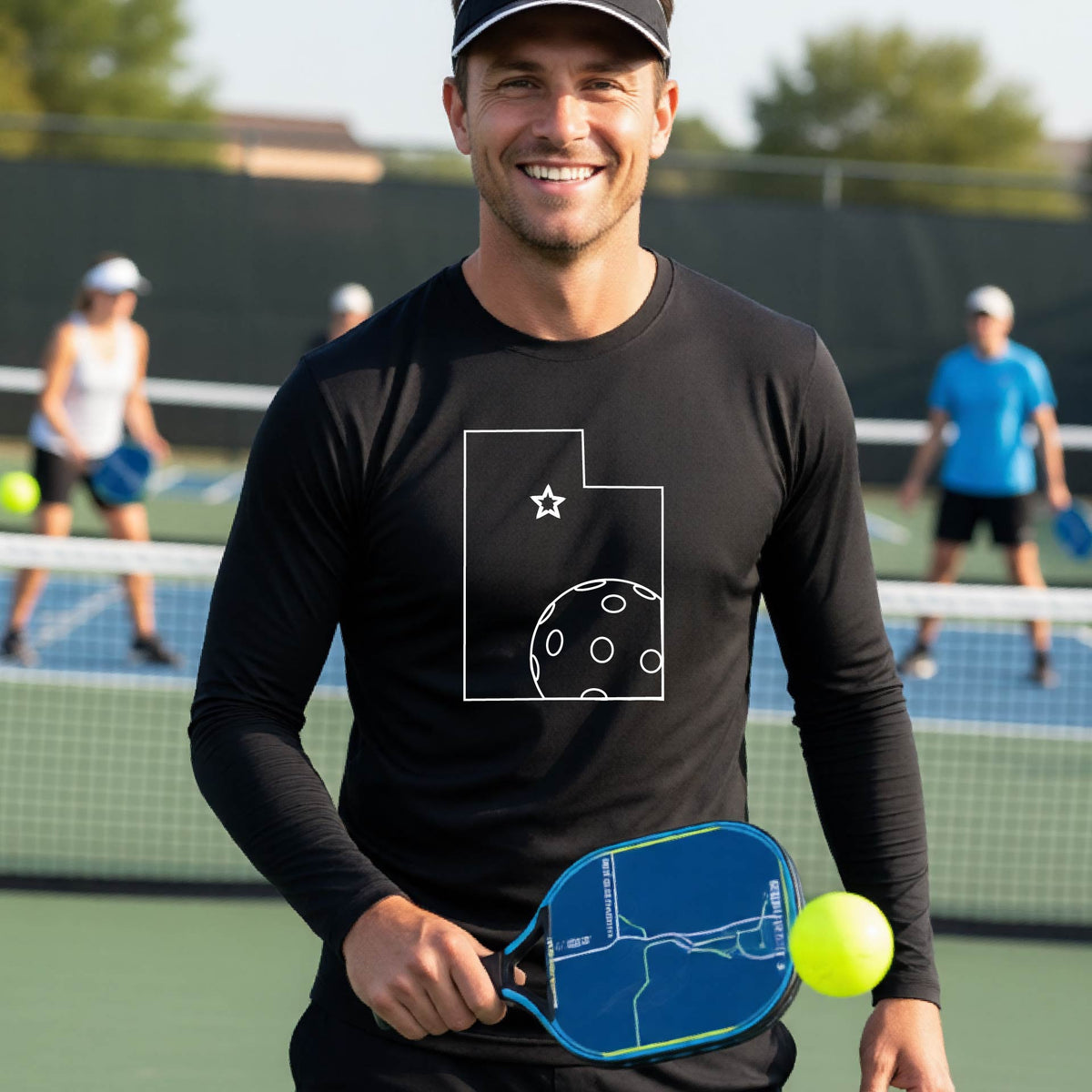 A close-up of a man wearing a black short-sleeve t-shirt, with a white graphic outline of the State of Utah , including an outline of a white pickleball in the lower right corner and a star outline in the center of the graphic, on the back of the shirt.   