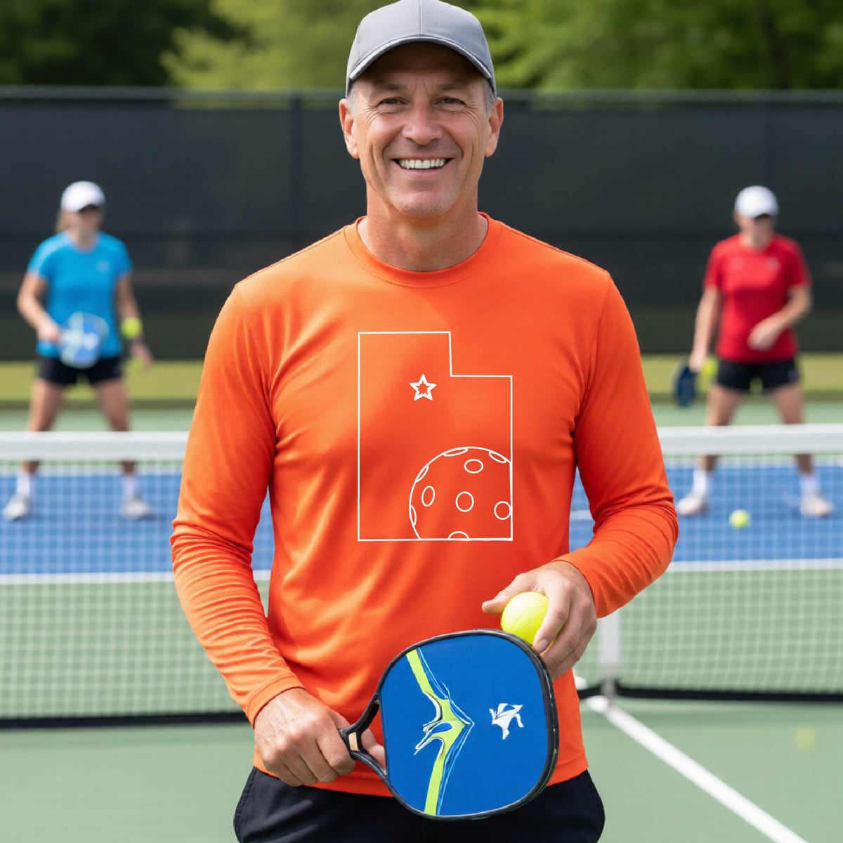A close-up of a man wearing an orange short-sleeve t-shirt, with a white graphic outline of the State of Utah , including an outline of a white pickleball in the lower right corner and a star outline in the center of the graphic, on the back of the shirt.   