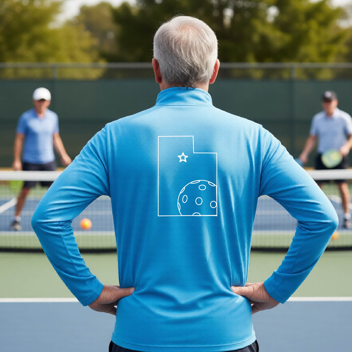 A close-up of a man, seen from behind, wearing a bright blue quarter-zip shirt, with a white graphic outline of the State of Utah , including an outline of a white pickleball in the lower right corner and a star outline in the center of the graphic, on the back of the shirt.   