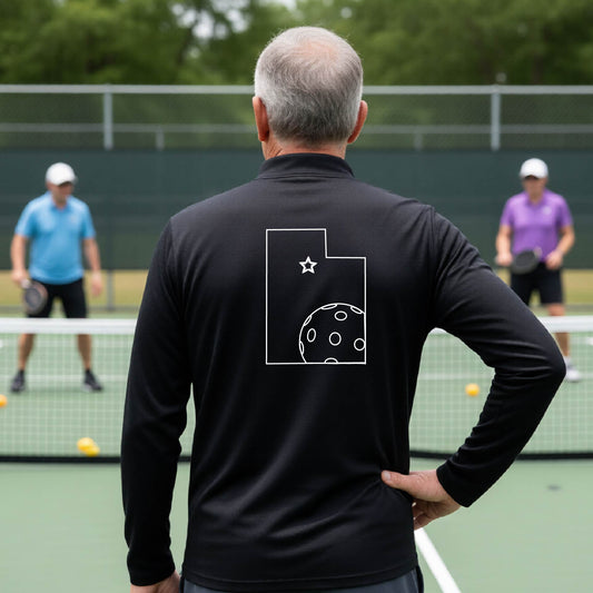 A close-up of a man, seen from behind, wearing a black quarter-zip shirt, with a white graphic outline of the State of Utah , including an outline of a white pickleball in the lower right corner and a star outline in the center of the graphic, on the back of the shirt.    