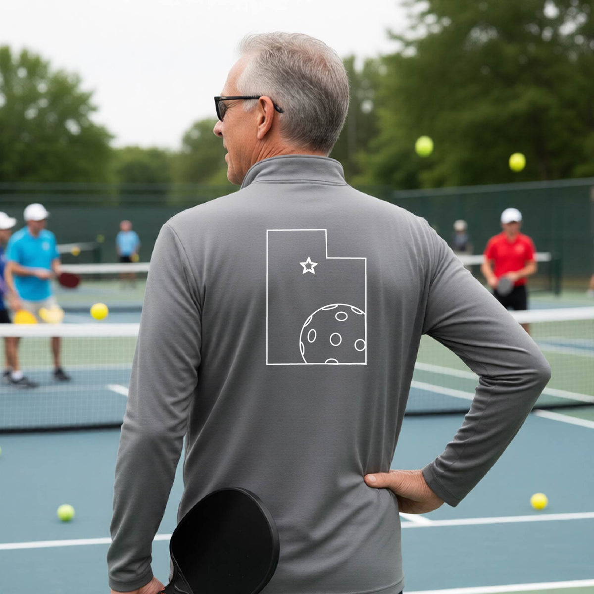 A close-up of a man, seen from behind, wearing a gray quarter-zip shirt, with a white graphic outline of the State of Utah , including an outline of a white pickleball in the lower right corner and a star outline in the center of the graphic, on the back of the shirt.   