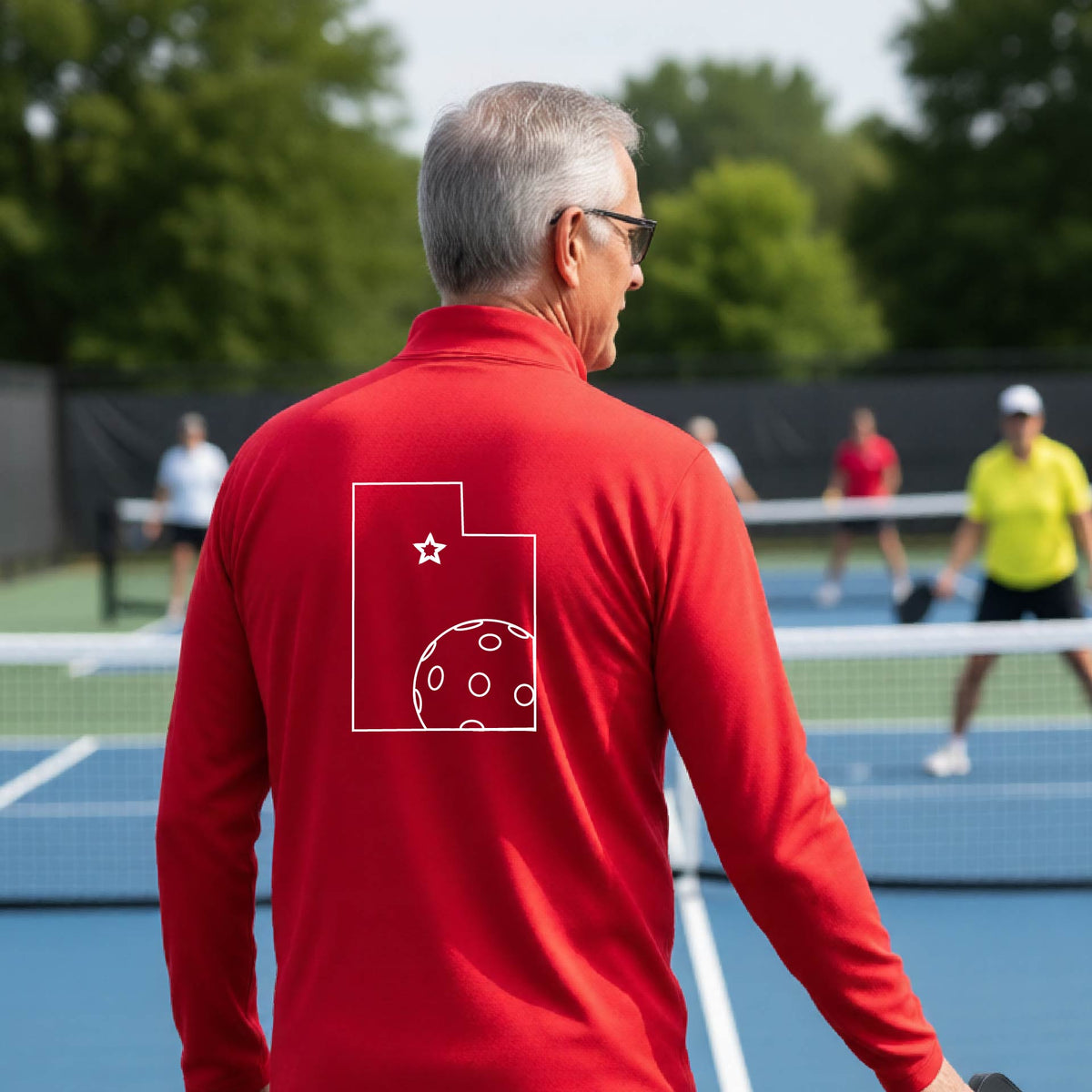 A close-up of a man, seen from behind, wearing a red quarter-zip shirt, with a white graphic outline of the State of Utah , including an outline of a white pickleball in the lower right corner and a star outline in the center of the graphic, on the back of the shirt.   
