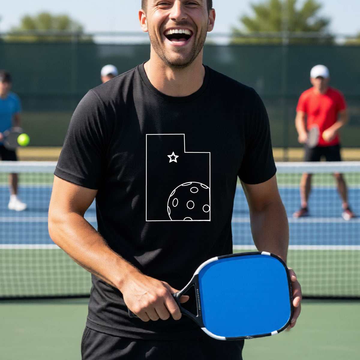 A close-up of a man wearing a black short-sleeve t-shirt, with a white graphic outline of the State of Utah , including an outline of a white pickleball in the lower right corner and a star outline in the center of the graphic, on the back of the shirt.   