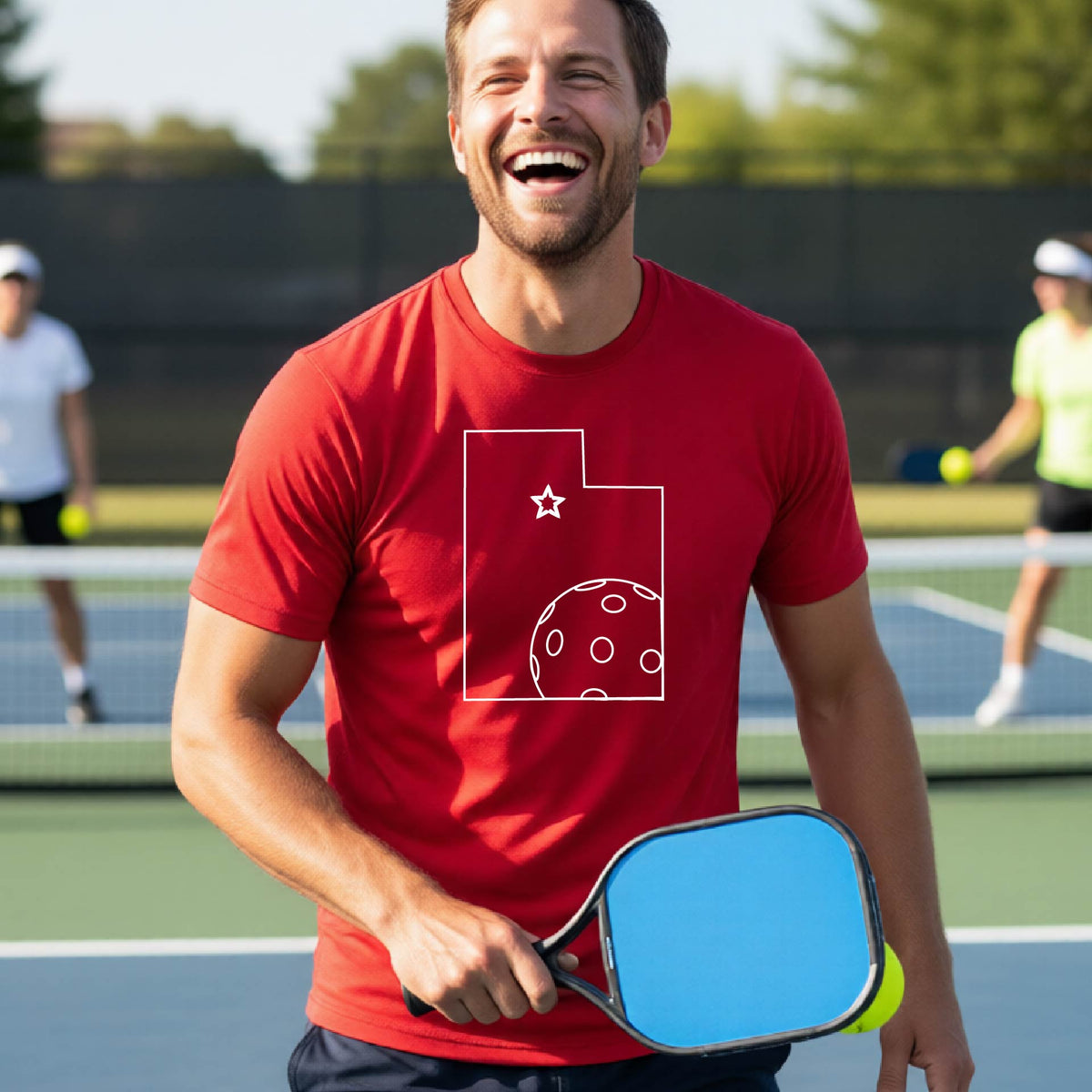 A close-up of a man wearing a red short-sleeve t-shirt, with a white graphic outline of the State of Utah , including an outline of a white pickleball in the lower right corner and a star outline in the center of the graphic, on the back of the shirt.   