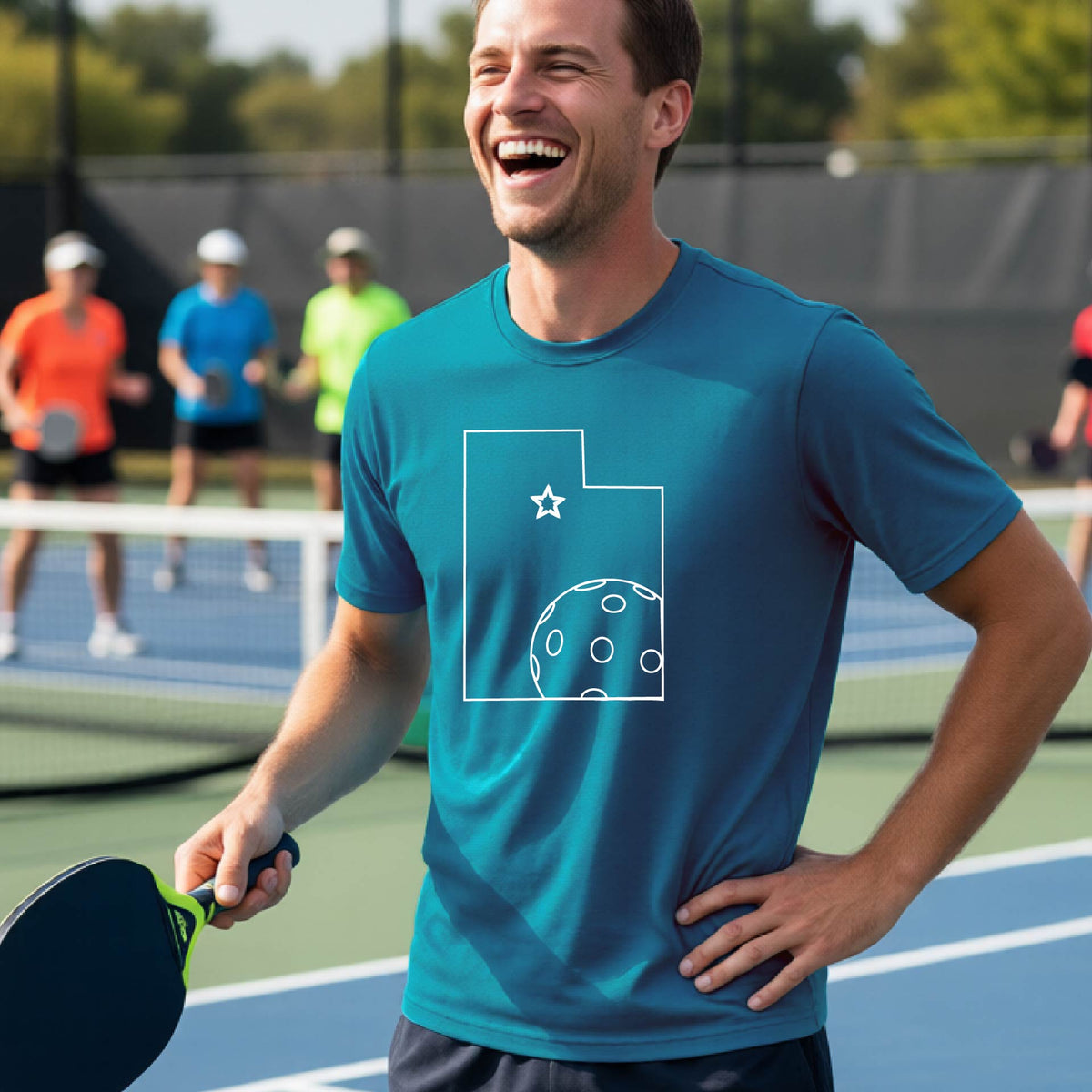 A close-up of a man wearing a teal short-sleeve t-shirt, with a white graphic outline of the State of Utah , including an outline of a white pickleball in the lower right corner and a star outline in the center of the graphic, on the back of the shirt.   