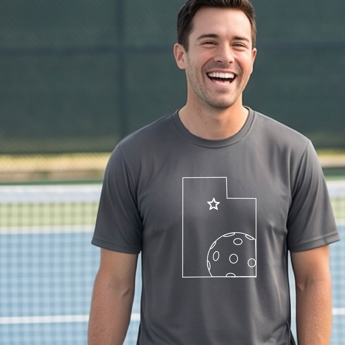 A close-up of a man wearing a dark gray short-sleeve t-shirt, with a white graphic outline of the State of Utah , including an outline of a white pickleball in the lower right corner and a star outline in the center of the graphic, on the back of the shirt.   