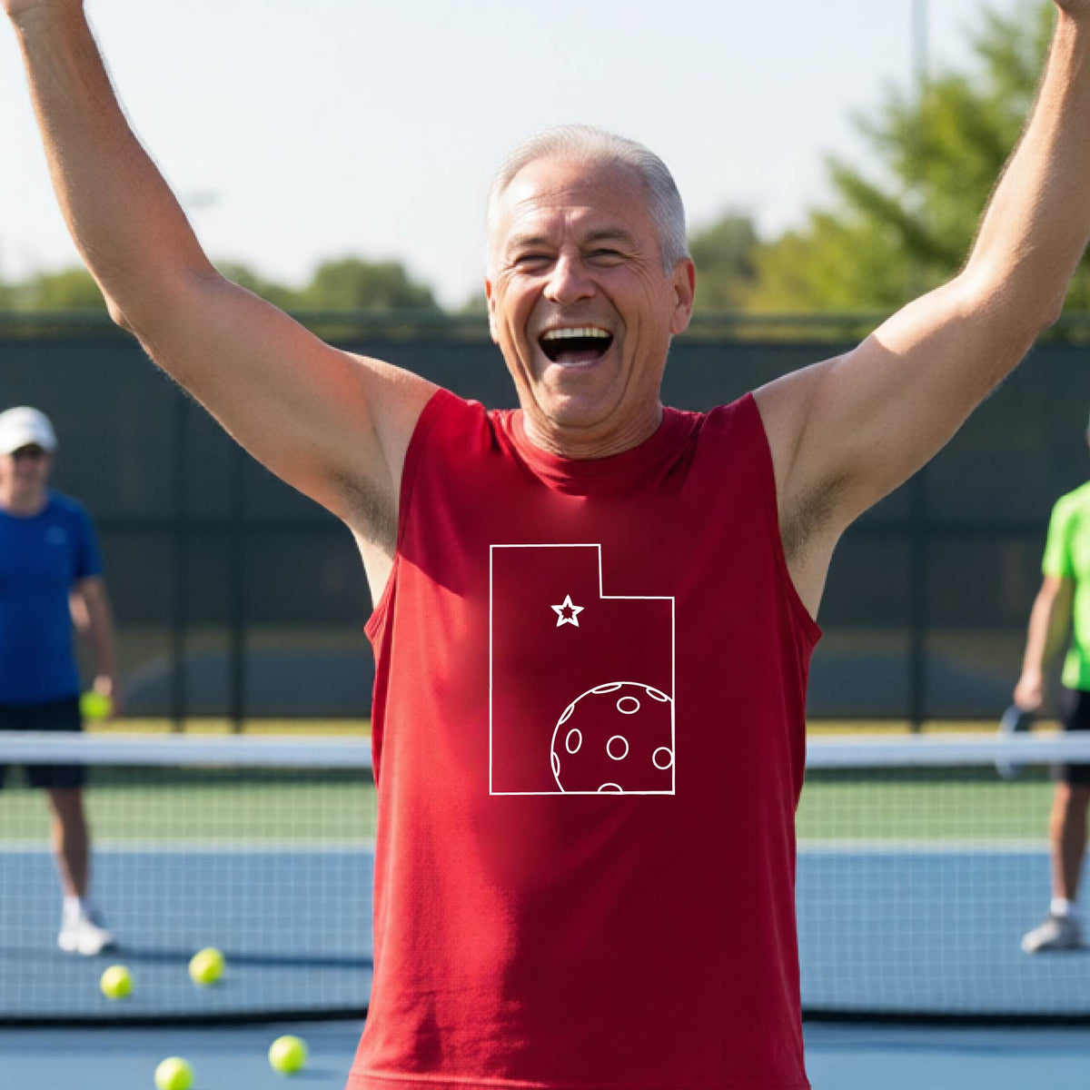 A close-up of a man wearing a red sleeveless t-shirt, with a white graphic outline of the State of Utah , including an outline of a white pickleball in the lower right corner and a star outline in the center of the graphic, on the back of the shirt.  