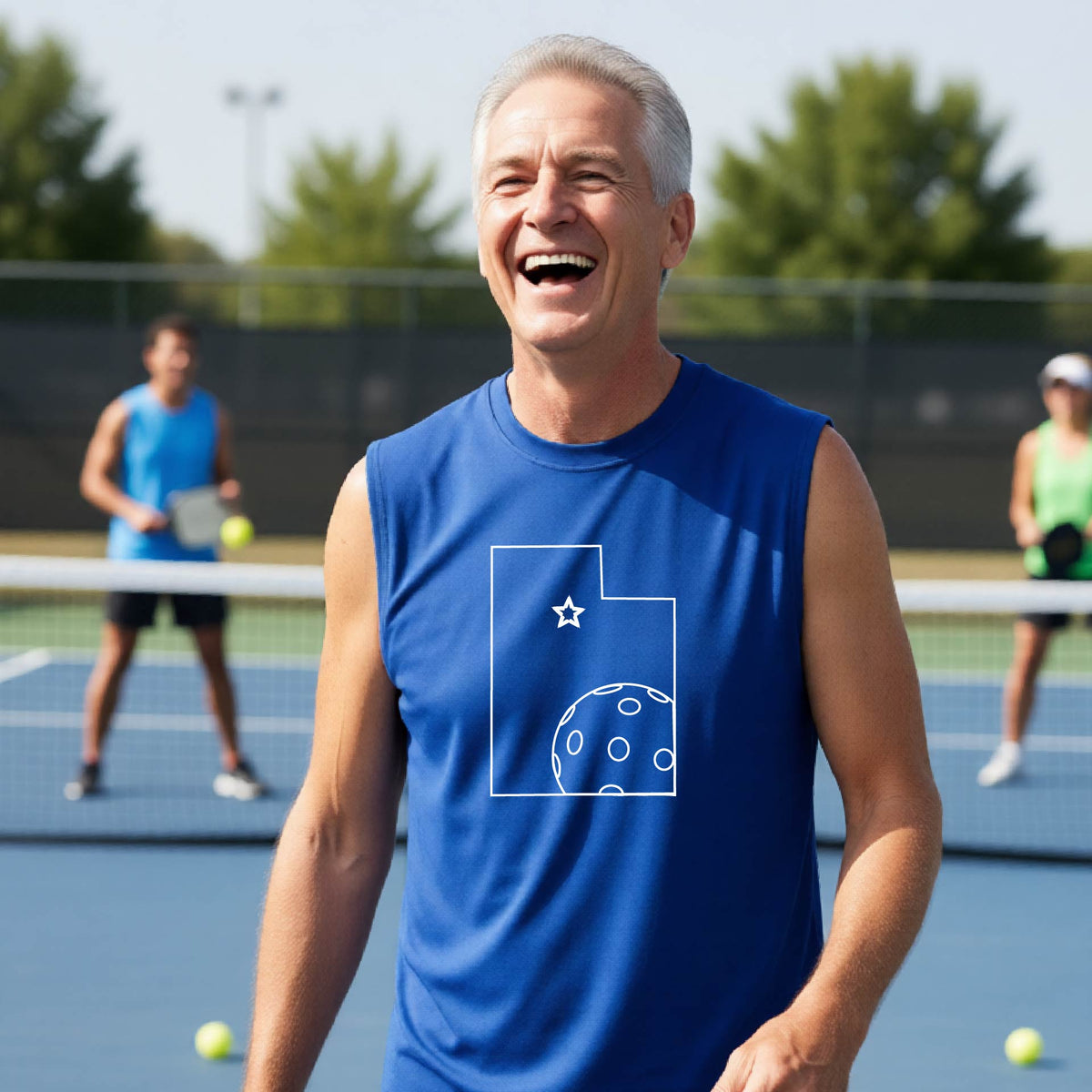 A close-up of a man wearing a royal blue sleeveless t-shirt, with a white graphic outline of the State of Utah , including an outline of a white pickleball in the lower right corner and a star outline in the center of the graphic, on the back of the shirt.  