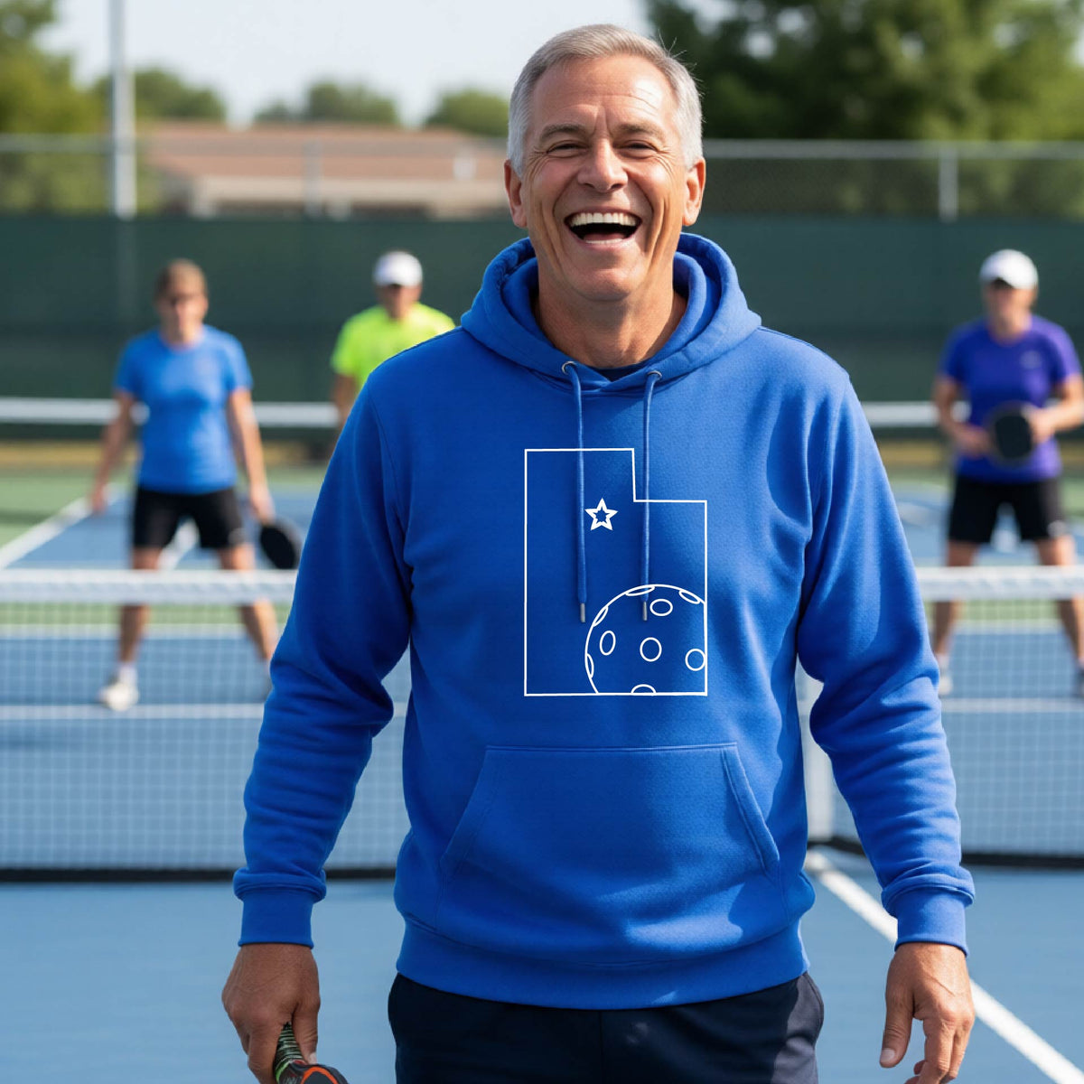 A close-up of a man wearing a royal blue unisex hoodie pullover, with a white graphic outline of the State of Utah , including an outline of a white pickleball in the lower right corner and a star outline in the center of the graphic, on the front of the hoodie. 