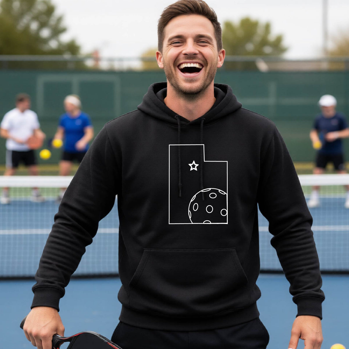 A close-up of a man wearing a black unisex hoodie pullover, with a white graphic outline of the State of Utah , including an outline of a white pickleball in the lower right corner and a star outline in the center of the graphic, on the front of the hoodie. 