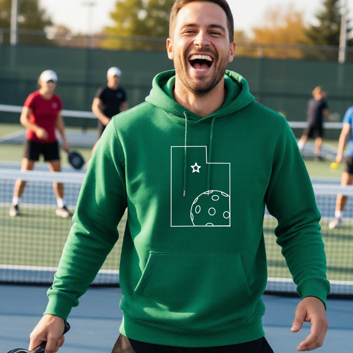 A close-up of a man wearing a green unisex hoodie pullover, with a white graphic outline of the State of Utah , including an outline of a white pickleball in the lower right corner and a star outline in the center of the graphic, on the front of the hoodie. 