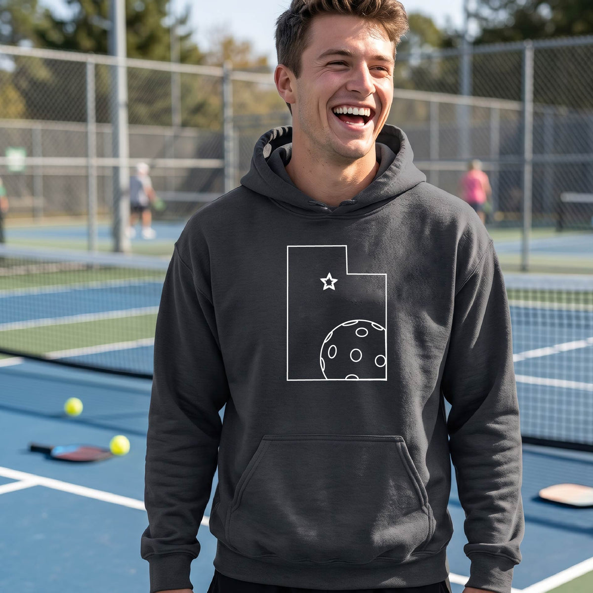 A close-up of a man wearing a dark gray unisex hoodie pullover, with a white graphic outline of the State of Utah , including an outline of a white pickleball in the lower right corner and a star outline in the center of the graphic, on the front of the hoodie. 