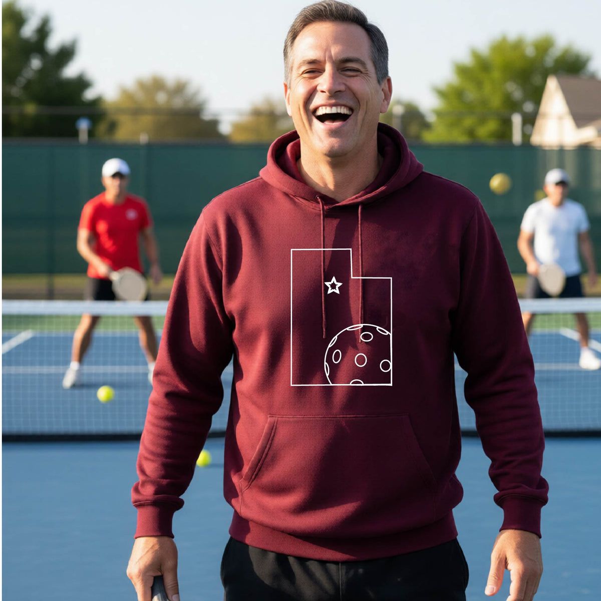 A close-up of a man wearing a maroon unisex hoodie pullover, with a white graphic outline of the State of Utah , including an outline of a white pickleball in the lower right corner and a star outline in the center of the graphic, on the front of the hoodie. 