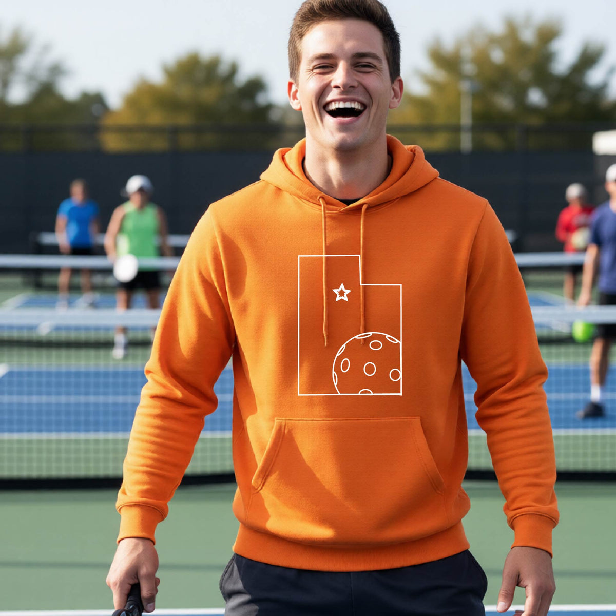 A close-up of a man wearing a black unisex hoodie pullover, with a white graphic outline of the State of Utah , including an outline of a white pickleball in the lower right corner and a star outline in the center of the graphic, on the front of the hoodie.   