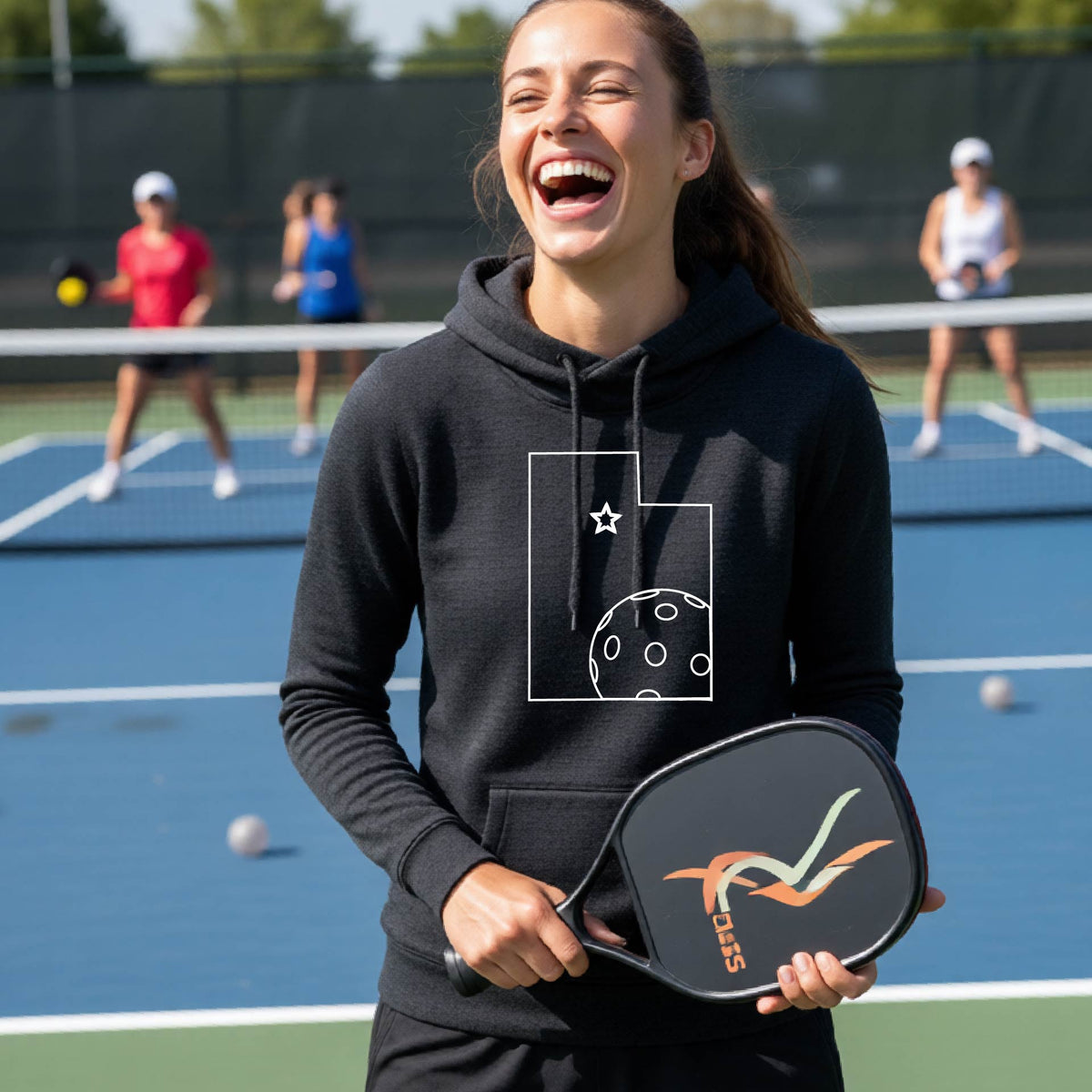 A close-up of a woman wearing a black cowl-neck hoodie pullover, with a white graphic outline of the State of Utah , including an outline of a white pickleball in the lower right corner and a star outline in the center of the graphic, on the front of the hoodie.   