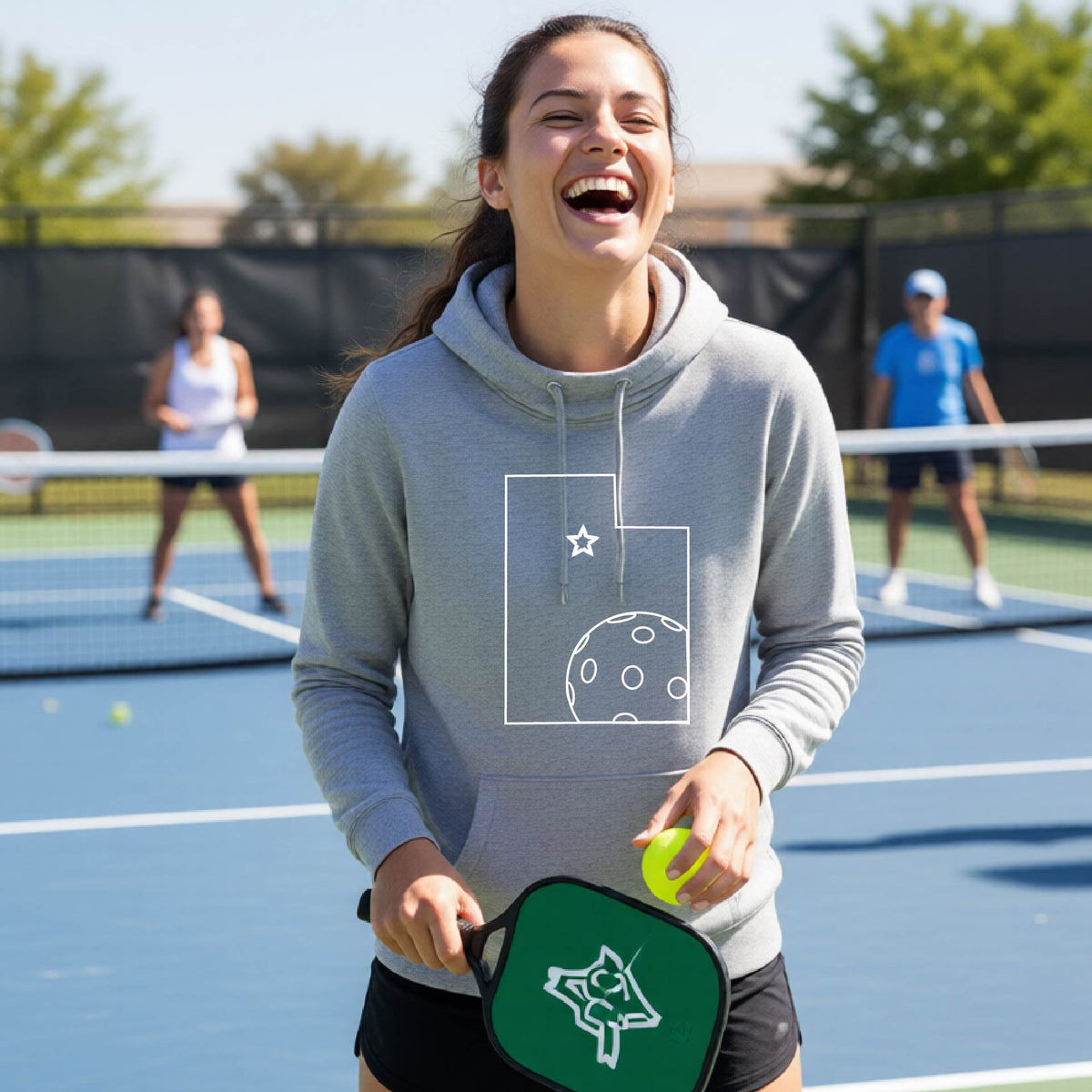 A close-up of a woman wearing a light gray cowl-neck hoodie pullover, with a white graphic outline of the State of Utah , including an outline of a white pickleball in the lower right corner and a star outline in the center of the graphic, on the front of the hoodie.   