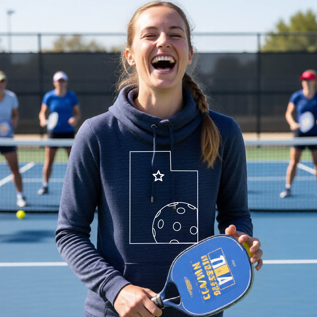 A close-up of a woman wearing a navy blue cowl-neck hoodie pullover, with a white graphic outline of the State of Utah , including an outline of a white pickleball in the lower right corner and a star outline in the center of the graphic, on the front of the hoodie.   