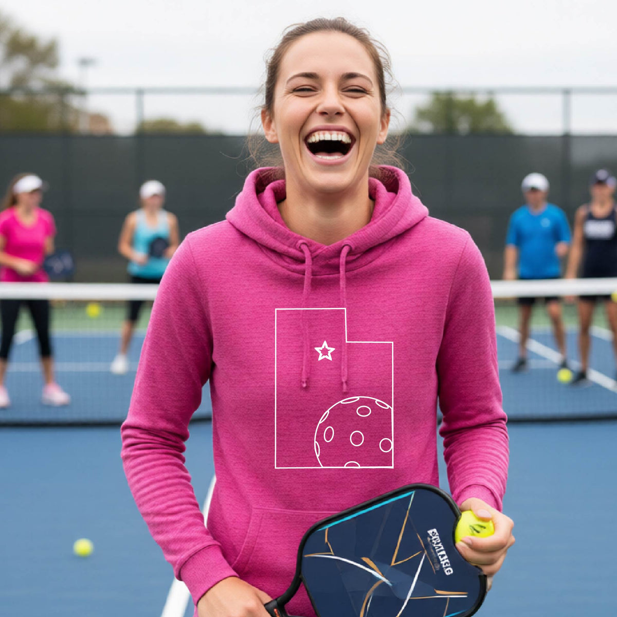 A close-up of a woman wearing a pink cowl-neck hoodie pullover, with a white graphic outline of the State of Utah , including an outline of a white pickleball in the lower right corner and a star outline in the center of the graphic, on the front of the hoodie.   