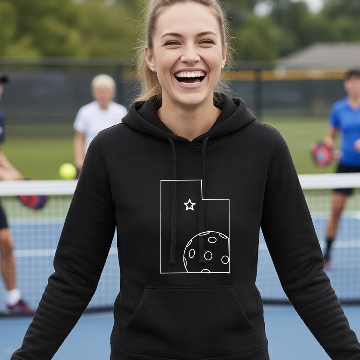 A close-up of a woman wearing a black fitted hoodie pullover, with a white graphic outline of the State of Utah , including an outline of a white pickleball in the lower right corner and a star outline in the center of the graphic, on the front of the hoodie.   