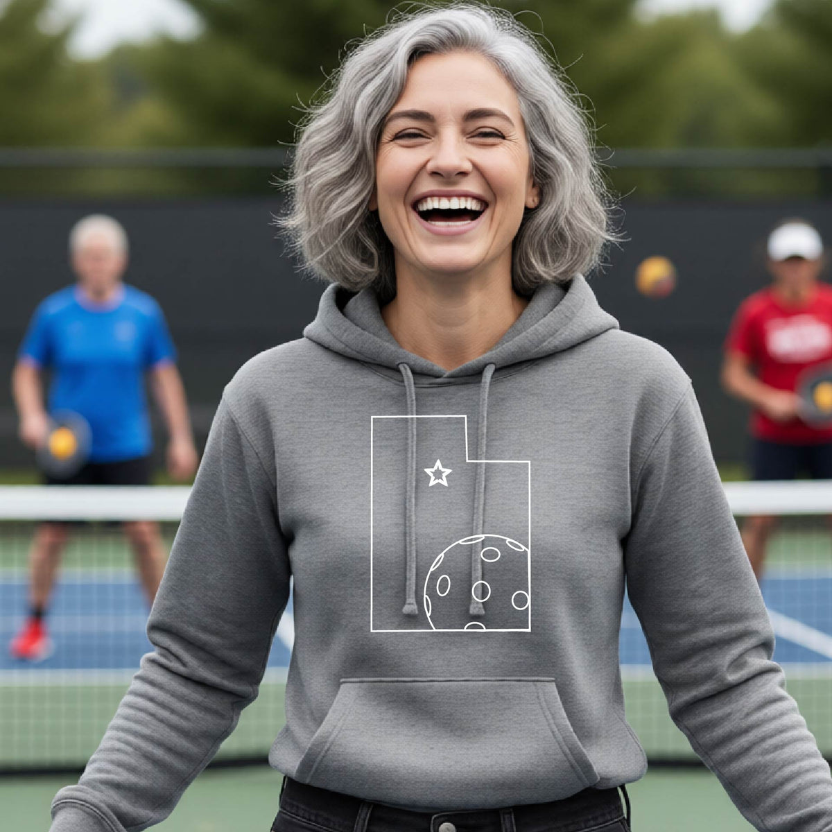 A close-up of a woman wearing a gray fitted hoodie pullover, with a white graphic outline of the State of Utah , including an outline of a white pickleball in the lower right corner and a star outline in the center of the graphic, on the front of the hoodie.   