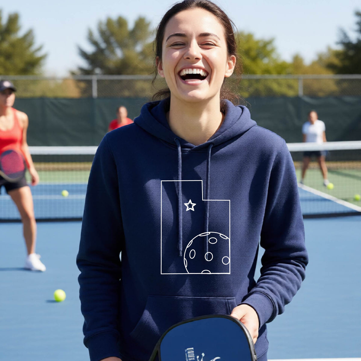 A close-up of a woman wearing a navy blue fitted hoodie pullover, with a white graphic outline of the State of Utah , including an outline of a white pickleball in the lower right corner and a star outline in the center of the graphic, on the front of the hoodie.   