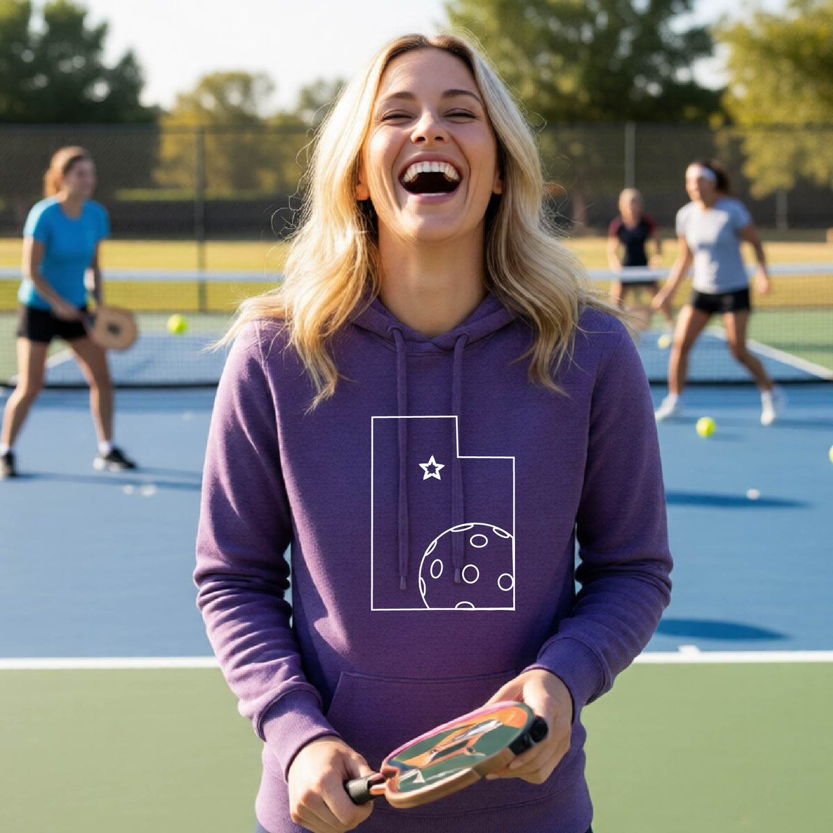 A close-up of a woman wearing a purple fitted hoodie pullover, with a white graphic outline of the State of Utah , including an outline of a white pickleball in the lower right corner and a star outline in the center of the graphic, on the front of the hoodie.   
