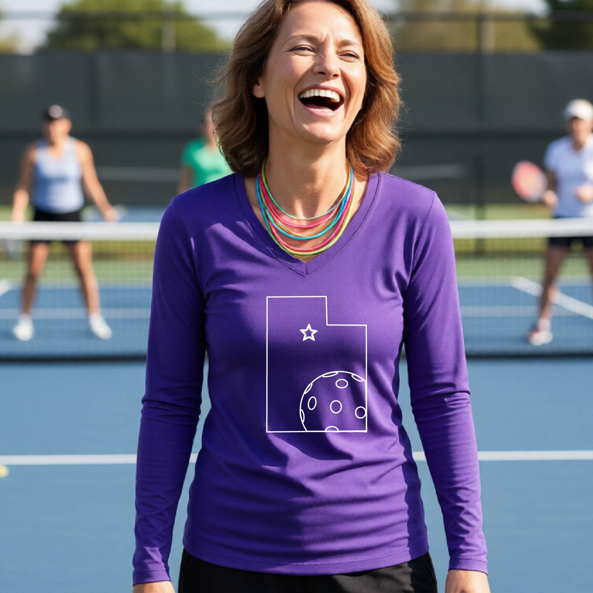 A close-up of a woman wearing a purple long-sleeve, with a white graphic outline of the State of Utah , including an outline of a white pickleball in the lower right corner and a star outline in the center of the graphic, on the back of the shirt.   