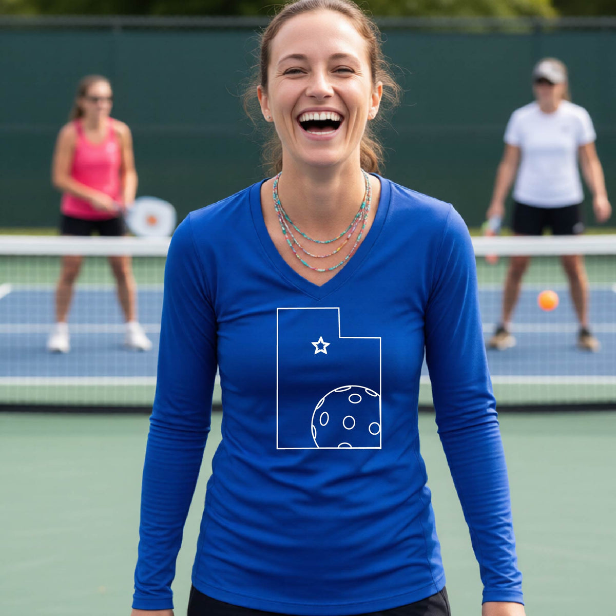 A close-up of a woman wearing a royal blue long-sleeve, with a white graphic outline of the State of Utah , including an outline of a white pickleball in the lower right corner and a star outline in the center of the graphic, on the back of the shirt.   