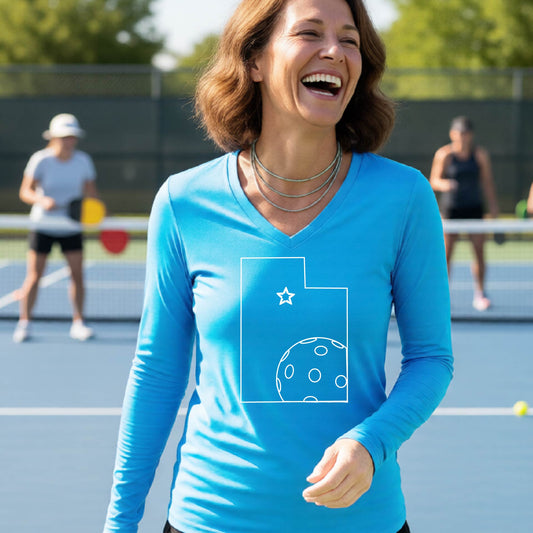 A close-up of a woman wearing a bright blue long-sleeve, with a white graphic outline of the State of Utah , including an outline of a white pickleball in the lower right corner and a star outline in the center of the graphic, on the back of the shirt.   