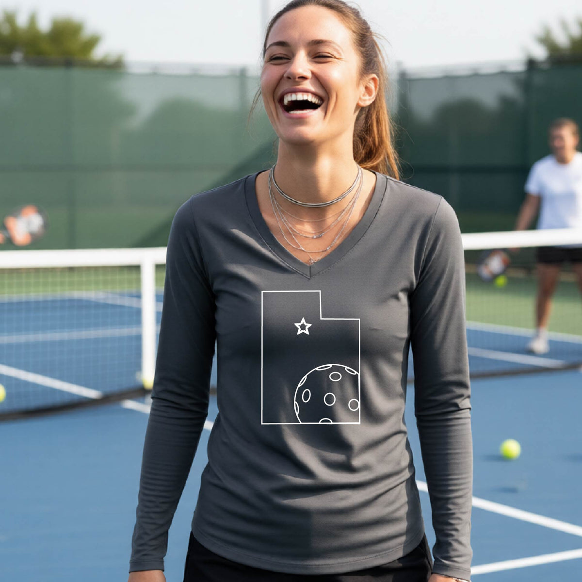 A close-up of a woman wearing a dark gray long-sleeve, with a white graphic outline of the State of Utah , including an outline of a white pickleball in the lower right corner and a star outline in the center of the graphic, on the back of the shirt.   