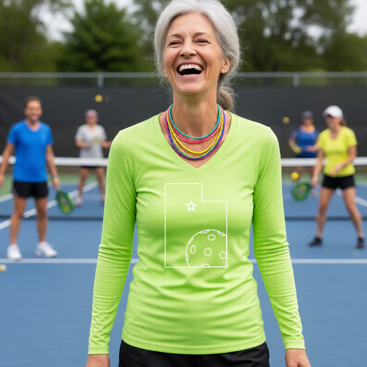 A close-up of a woman wearing a lime green long-sleeve, with a white graphic outline of the State of Utah , including an outline of a white pickleball in the lower right corner and a star outline in the center of the graphic, on the back of the shirt.   