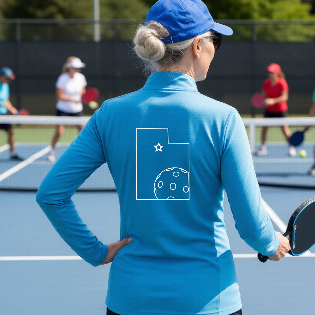 A close-up of a woman, seen from behind, wearing a bright blue quarter-zip shirt, with a white graphic outline of the State of Utah , including an outline of a white pickleball in the lower right corner and a star outline in the center of the graphic, on the back of the shirt.   