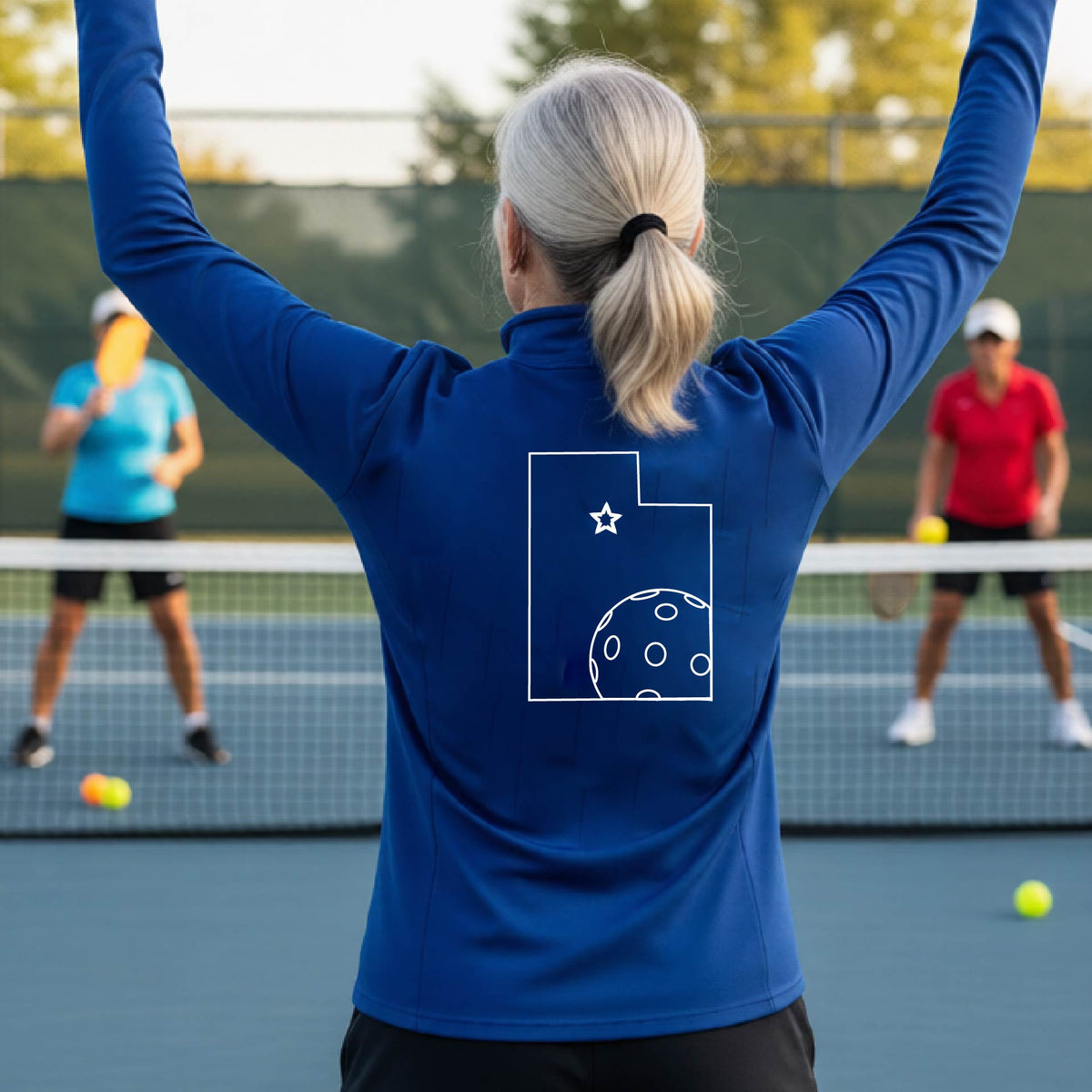 A close-up of a woman, seen from behind, wearing a royal blue quarter-zip shirt, with a white graphic outline of the State of Utah , including an outline of a white pickleball in the lower right corner and a star outline in the center of the graphic, on the back of the shirt.   
