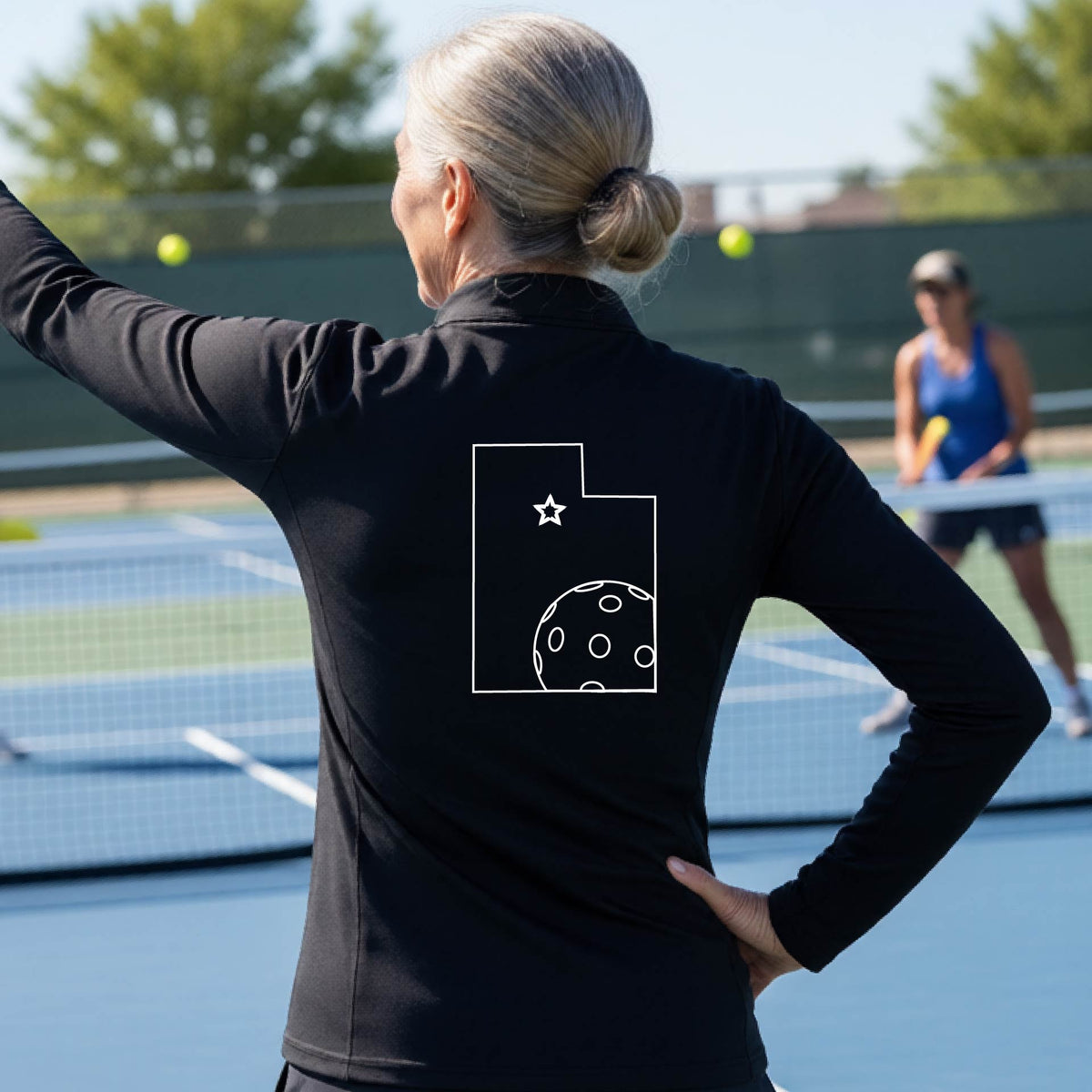 A close-up of a woman, seen from behind, wearing a black quarter-zip shirt, with a white graphic outline of the State of Utah , including an outline of a white pickleball in the lower right corner and a star outline in the center of the graphic, on the back of the shirt.   