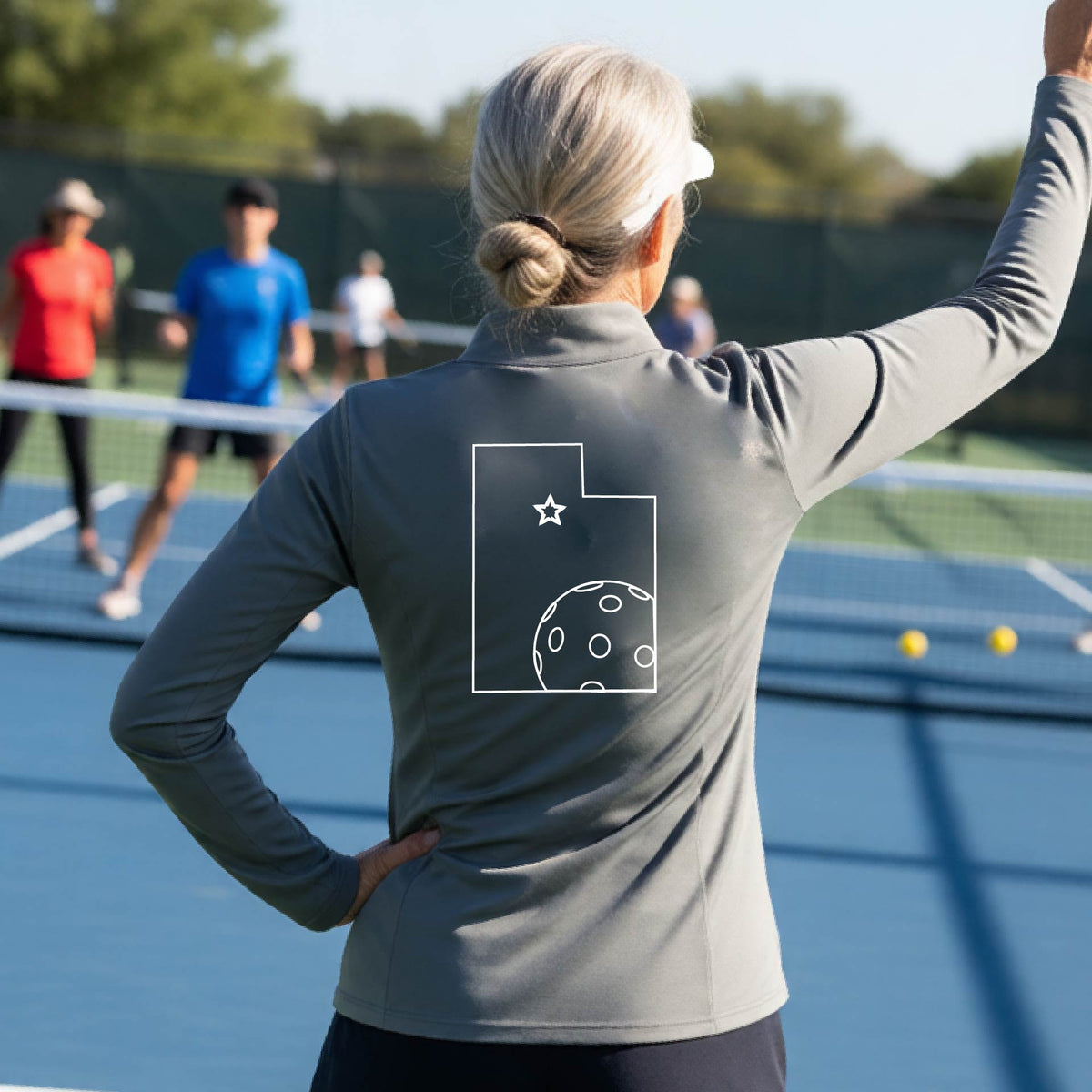 A close-up of a woman, seen from behind, wearing a gray quarter-zip shirt, with a white graphic outline of the State of Utah , including an outline of a white pickleball in the lower right corner and a star outline in the center of the graphic, on the back of the shirt.   
