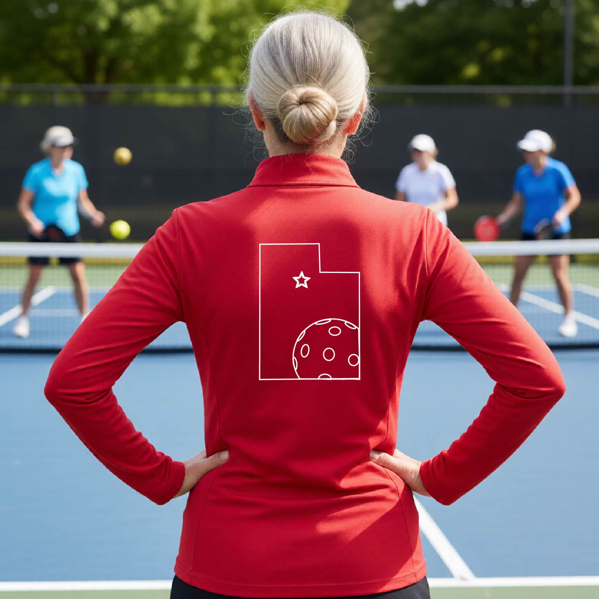 A close-up of a woman, seen from behind, wearing a red quarter-zip shirt, with a white graphic outline of the State of Utah , including an outline of a white pickleball in the lower right corner and a star outline in the center of the graphic, on the back of the shirt.   
